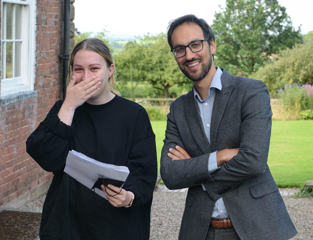 A happy girl smiling at her A Level results with a teacher stood next to her